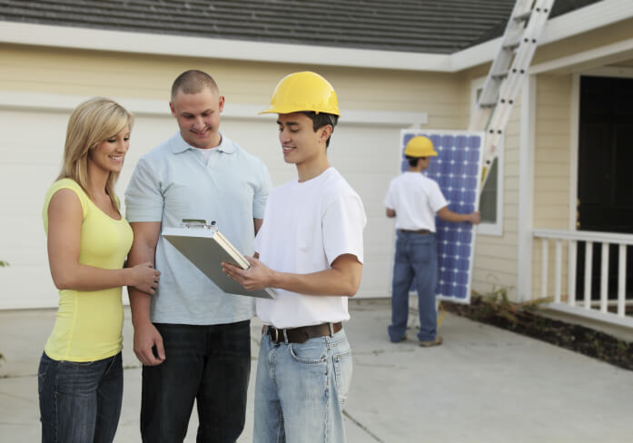 Contractor speaking with homeowners. Contract holding a solar panel in the background.