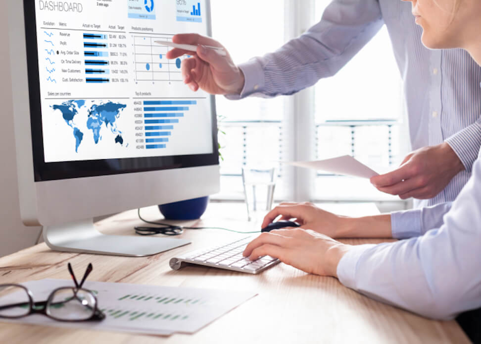 One person pointing to a chart on a computer screen. A second person typing on a keyboard.
